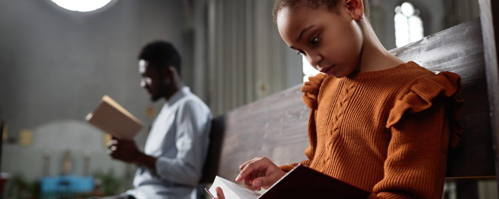 African American little girl reading Bible while sitting on bench in church with man in background